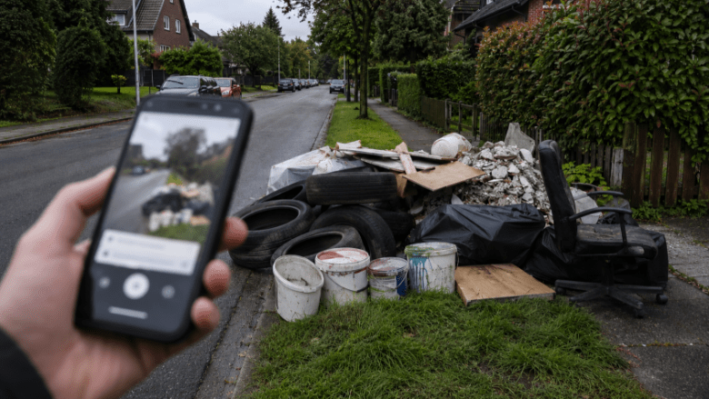 Ein Smartphone fotografiert eine illegale Müllablagerung an einer Wohnstraße. Im Hintergrund liegen alte Reifen, Farbeimer und Müllsäcke am Straßenrand vor typischen norddeutschen Wohnhäusern.